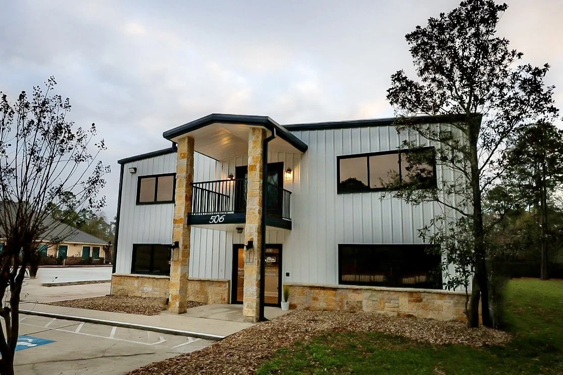 The clinic building at dusk: a two-storey white metal and stone office with the street number 506 above the entrance and an accessible parking space in front.